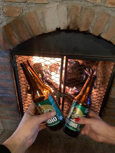 Dos manos sosteniendo botellas de cerveza frente a una chimenea encendida.