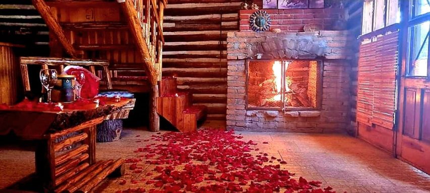 Interior de una cabaña con una chimenea encendida y pétalos de rosa en el suelo.