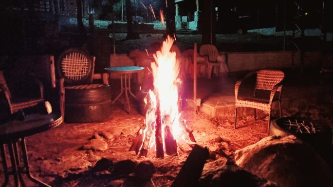 Fogata iluminando un entorno oscuro con sillas alrededor y un ambiente acogedor.
