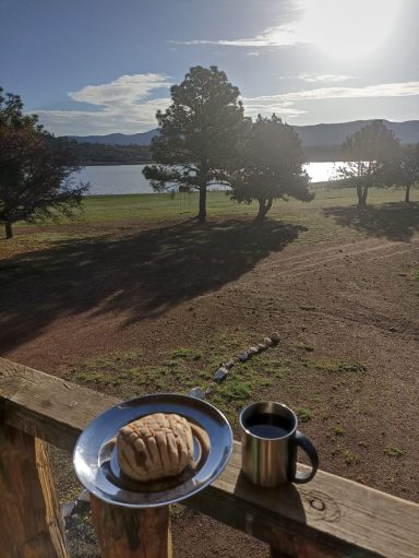 Plato con pan y taza de café frente a un lago, bajo un cielo soleado.